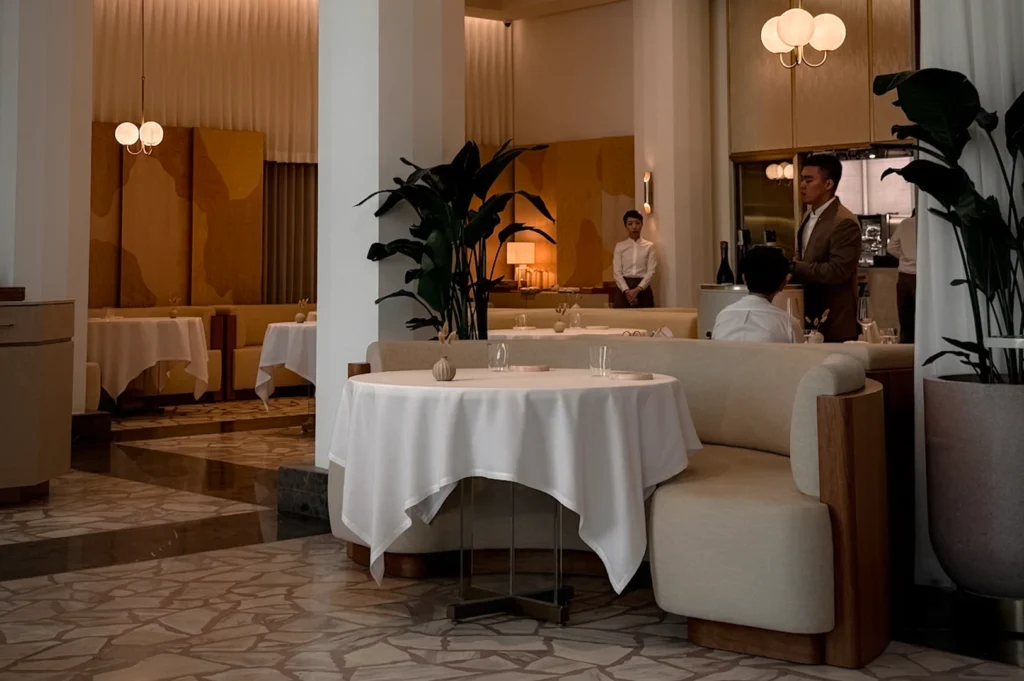 The elegant, minimalist interior of Odette restaurant at the National Gallery Singapore. The fine dining space features soft pastel tones, curved banquette seating, and crisp white tablecloths. Warm ambient lighting and lush indoor plants complement the sophisticated atmosphere of this Michelin-starred establishment.
