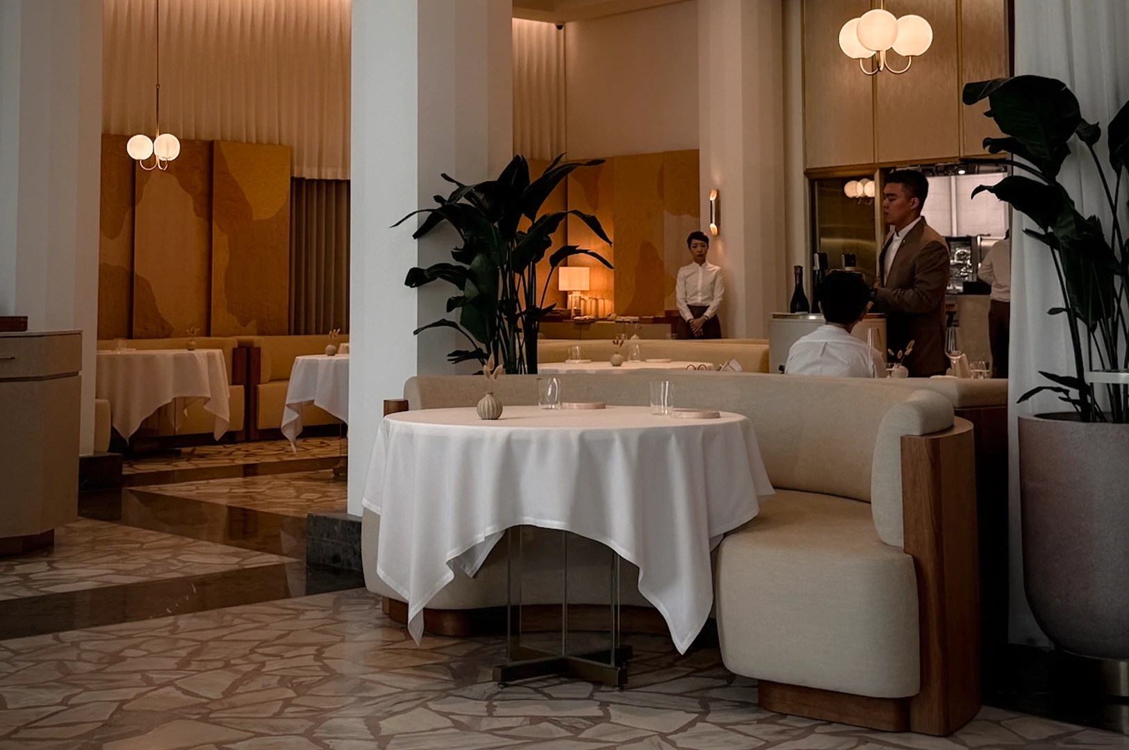 The elegant, minimalist interior of Odette restaurant at the National Gallery Singapore. The fine dining space features soft pastel tones, curved banquette seating, and crisp white tablecloths. Warm ambient lighting and lush indoor plants complement the sophisticated atmosphere of this Michelin-starred establishment.