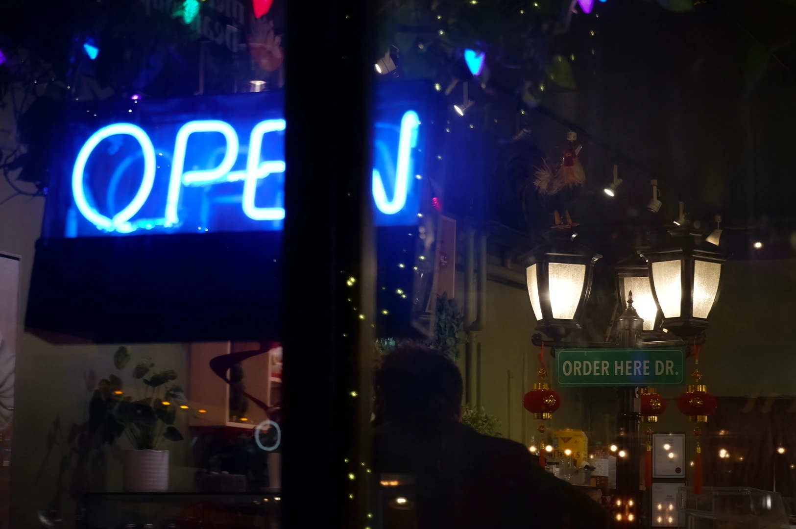Night view through a restaurant window with a glowing blue neon “OPEN” sign, warm street-style lantern lights, and a cozy dining interior decorated with plants and hanging ornaments.