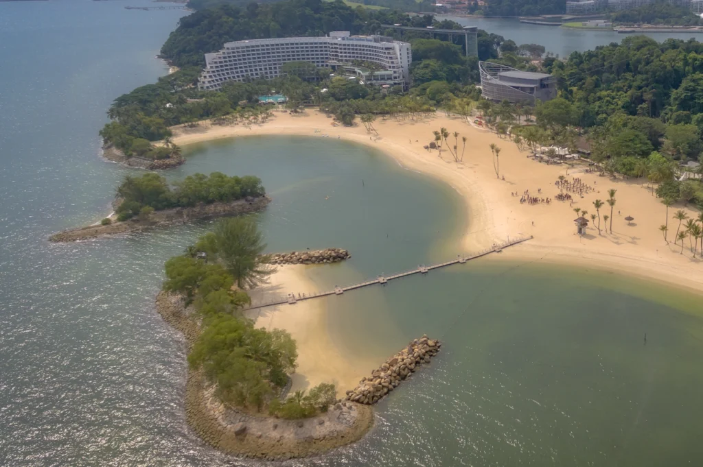 Aerial view of Shangri-La Rasa Sentosa in Singapore featuring a curved beachfront, calm lagoon waters, palm-lined sandy beach, and a large seaside resort surrounded by lush tropical greenery.
