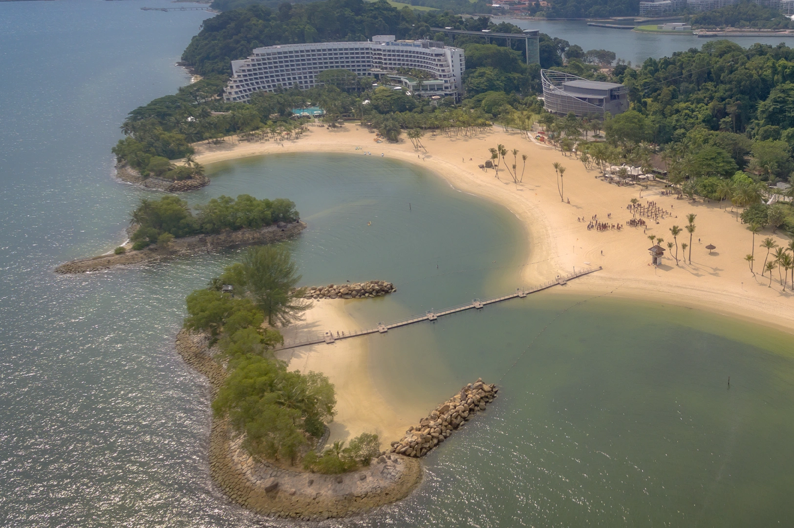 Aerial view of Shangri-La Rasa Sentosa in Singapore featuring a curved beachfront, calm lagoon waters, palm-lined sandy beach, and a large seaside resort surrounded by lush tropical greenery.
