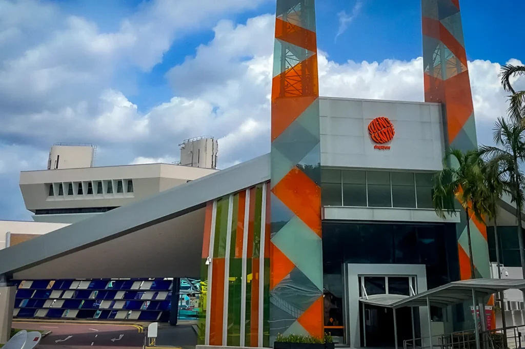 Exterior view of the Singapore Science Centre featuring its modern geometric facade with orange and green accents, tall entrance towers, and a cloudy blue sky in the background