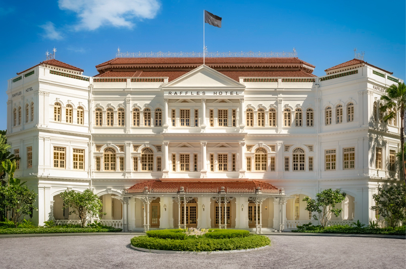 Luxury colonial-style facade of Raffles Hotel Singapore with white neoclassical architecture, red-tiled roofs, symmetrical windows, and landscaped front lawn under a clear blue sky.