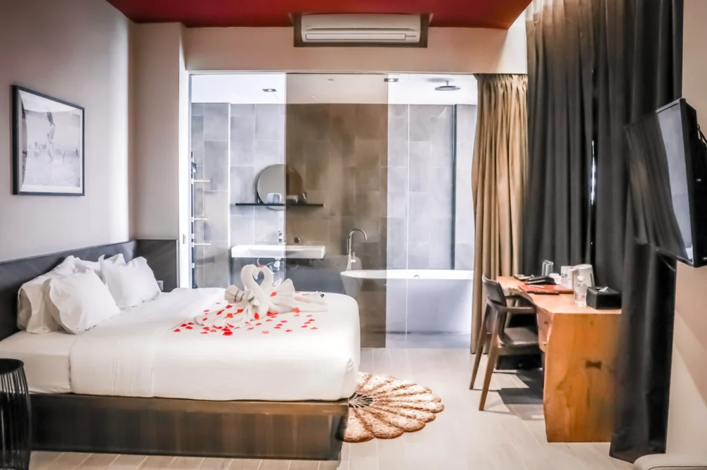  The romantic guest room interior at Hotel Yan in Singapore, featuring a king-sized bed decorated with swan-folded towels and red rose petals. The industrial-chic design includes a glass-walled bathroom with a standalone bathtub, a rustic wooden desk, and warm lighting. A wall-mounted TV and dark curtains complete the intimate and modern atmosphere.