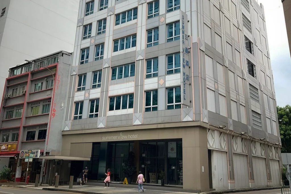 The exterior building view of Summer View Hotel in Singapore, featuring a modern multi-story facade with light grey paneling and blue-tinted windows. The hotel entrance is located at street level with large glass doors and a prominent sign. The surrounding urban area shows adjacent buildings and pedestrians on the sidewalk under a bright sky.