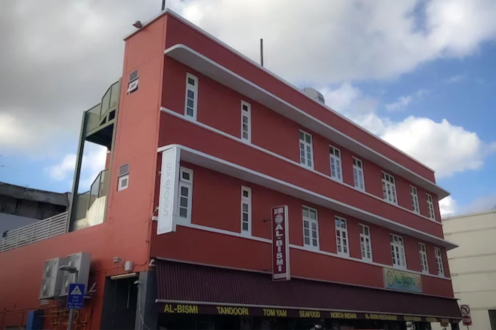 The exterior building view of Snooze Inn in Singapore, featuring a striking three-story facade painted in a vibrant terracotta red. The heritage-style building is characterized by its rows of classic white-framed windows and a prominent vertical "Snooze Inn" sign.