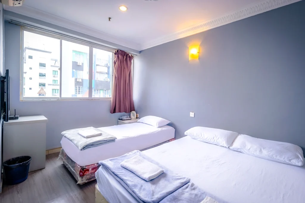 The interior view of a guest room at K Hotel Geylang in Singapore, featuring a simple and practical layout with two beds—a queen-sized bed and a single bed. Both beds are topped with white linens, light blue bed runners, and folded white towels. The room is painted in a soft greyish-blue hue, accented by a warm wall-mounted light and a large window that lets in natural light and offers a view of the surrounding urban buildings.