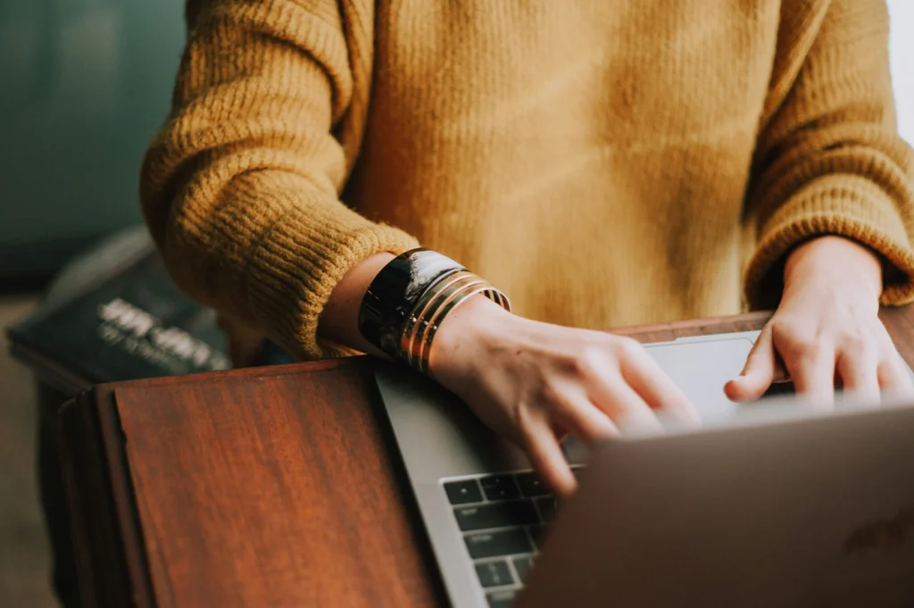 A person wearing a mustard yellow sweater and gold bracelets typing on a laptop at a wooden desk.