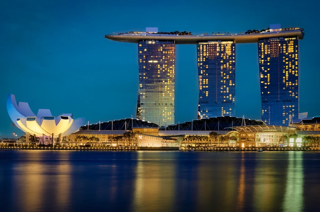 Night view of the Marina Bay Sands hotel in Singapore, featuring its three towers and the Sands SkyPark. The ArtScience Museum is illuminated in white, reflecting off the calm water of the bay alongside city lights.