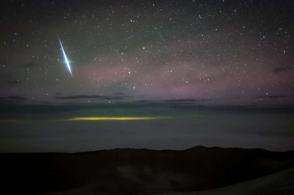 Bright Geminid meteor streaking across a star-filled night sky above dark landscape silhouettes and glowing clouds near the horizon