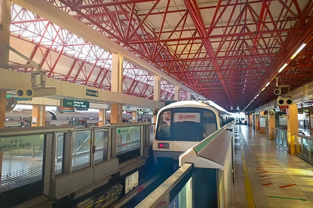 SMRT train arriving at Jurong East MRT Station in Singapore with red steel roof trusses, platform barriers, and bright natural light inside the station platform area