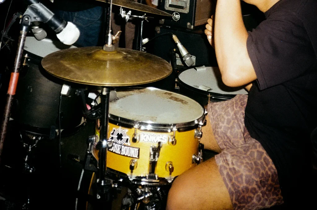 Drummer performing during a live indie music concert with band members, drum kit and stage equipment in a dimly lit venue atmosphere