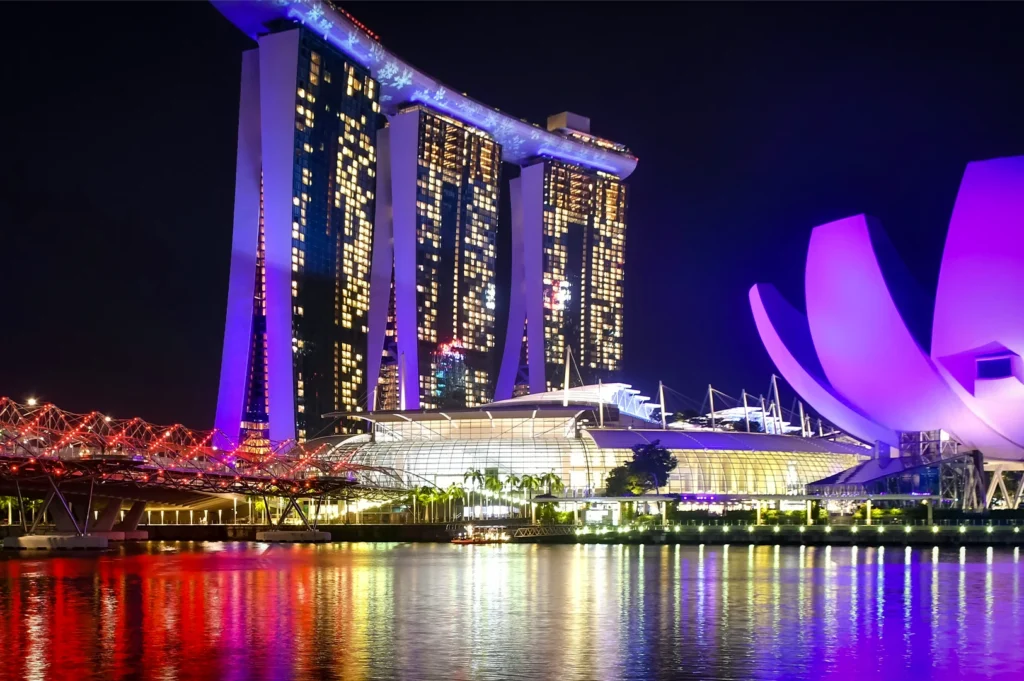 Marina Bay Sands and the ArtScience Museum illuminated at night in Singapore, with colorful reflections on the water and a lit pedestrian bridge in the foreground.