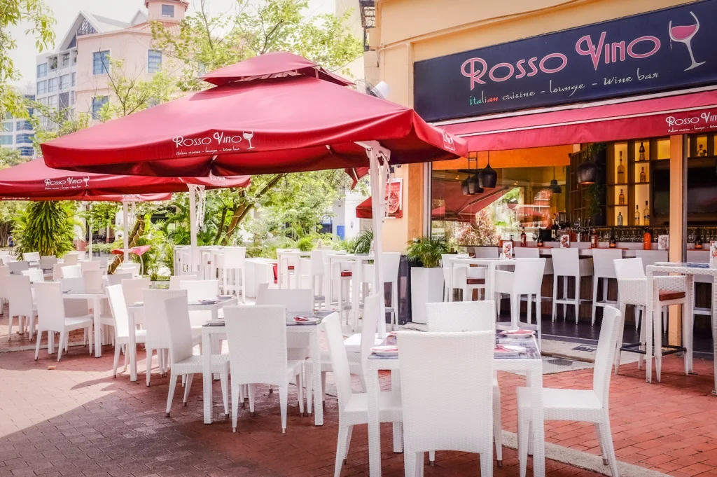 Outdoor seating area of Rosso Vino Italian restaurant with red umbrellas, white wicker chairs, and open bar, set along a sunny street with lush greenery and modern buildings in the background.