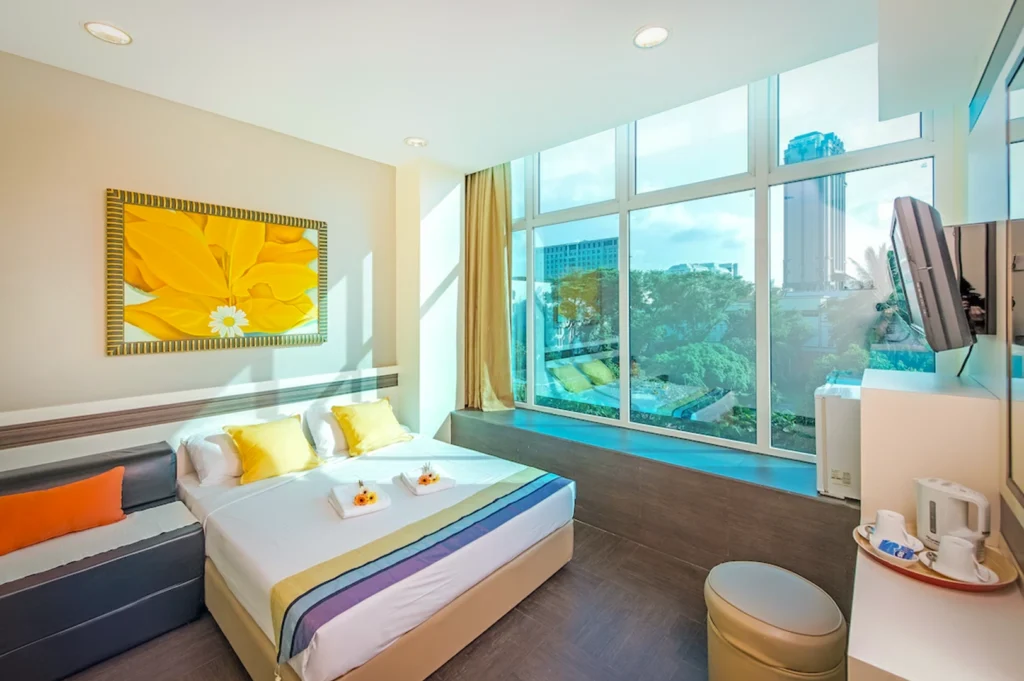 Interior of a modern guest room at Hotel 81 Bugis in Singapore. The room features a single bed with white linens, and a wall-mounted television. A window with yellow curtains provides a view of the surrounding urban buildings.