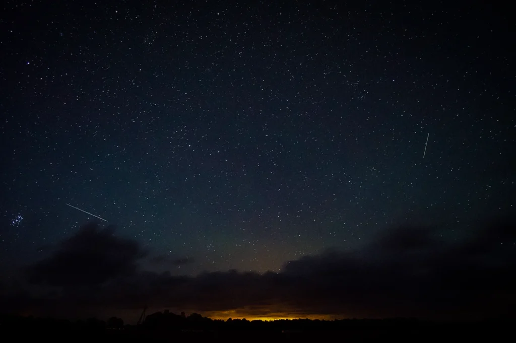 Meteor shower streaking across a star-filled night sky above dark clouds and a glowing horizon over a quiet landscape at night