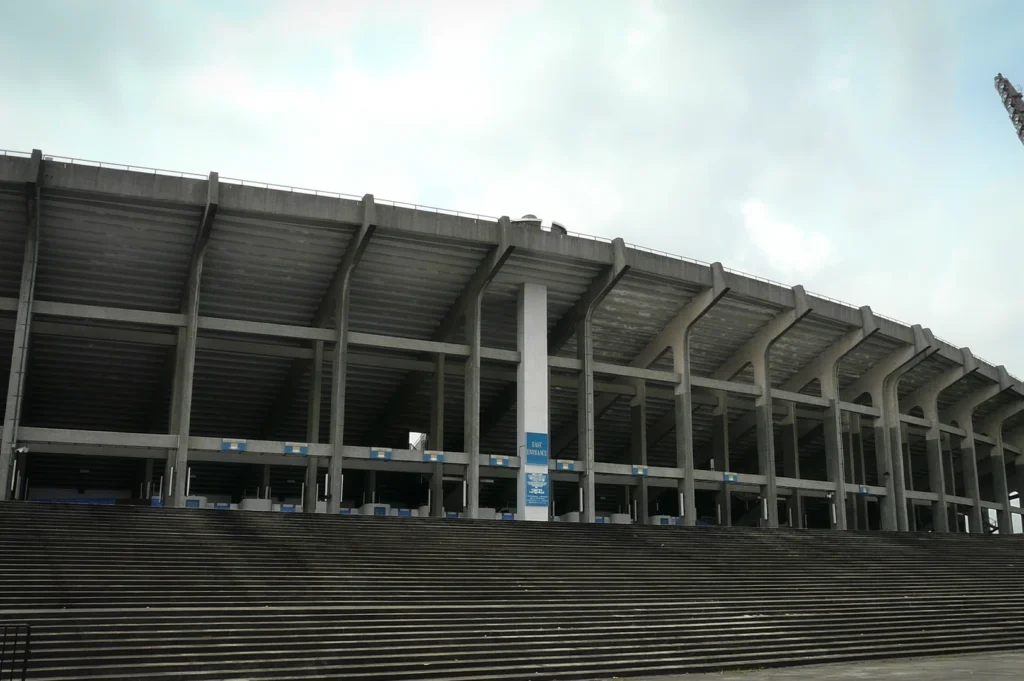 Exterior of the National Stadium in Kallang, Singapore, showing its concrete structure, wide steps, and tall floodlight tower under an overcast sky.