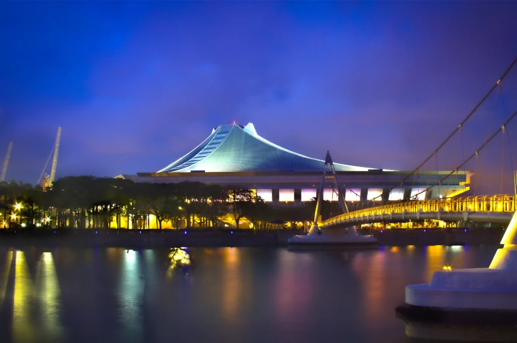 A futuristic building with a curved, tent-like roof is illuminated at night, reflected in the calm waters of a river, with a suspension bridge nearby.