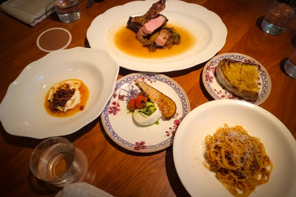 Overhead view of a gourmet Italian dining table featuring pasta, burrata, roasted meat, and artisan bread, beautifully plated on ceramic dishes in a warm, rustic restaurant setting.