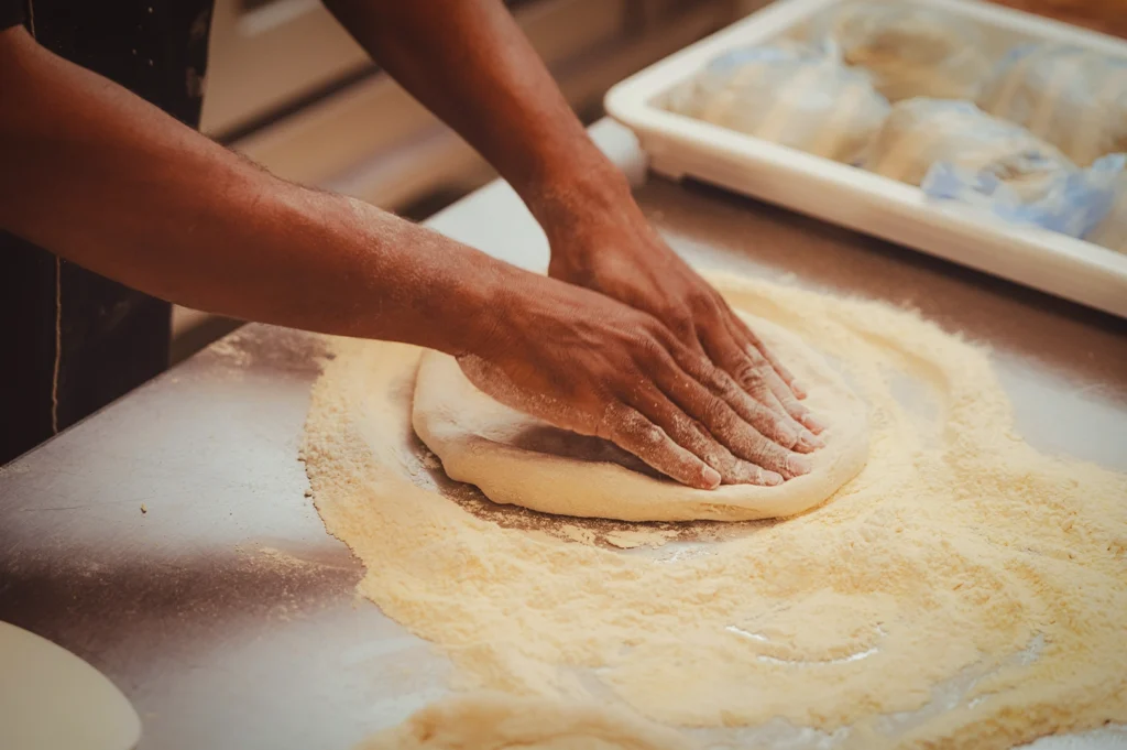 Chef kneading fresh dough on a floured metal table, preparing bread or pizza in a professional kitchen, showcasing hands-on baking process, texture, and artisanal food preparation.