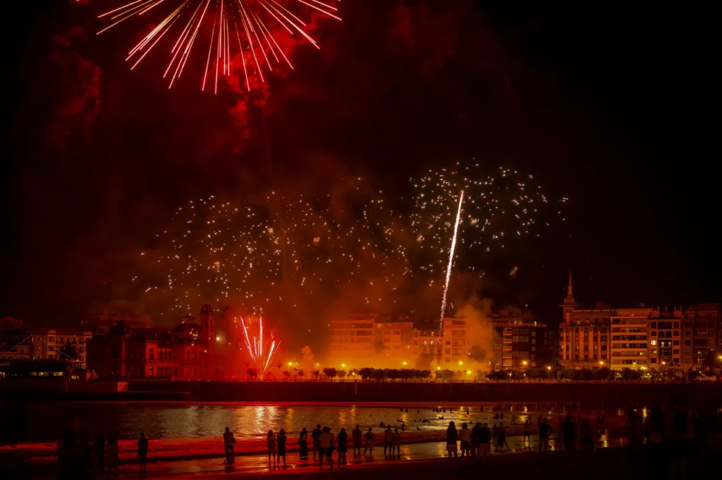 Colorful fireworks lighting up the night sky above a beachfront city with crowds watching from the shore and reflections glowing on the water