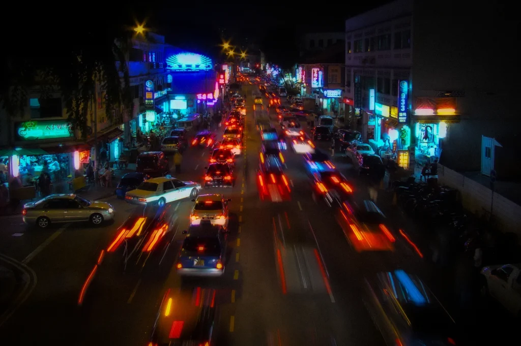 Busy nighttime street scene in Geylang district Singapore featuring neon signs, heavy traffic, colorful city lights, and long exposure light trails along Geylang Road