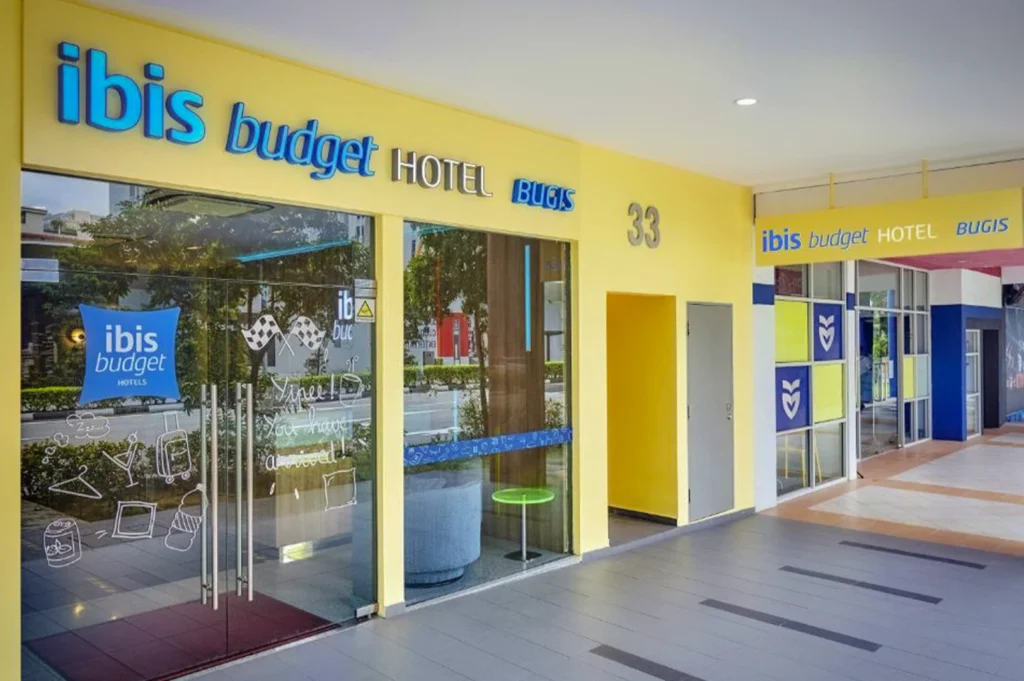 Front view of ibis budget Hotel Bugis in Singapore, featuring bright yellow facade, glass entrance doors, blue signage, and modern exterior design along a clean urban sidewalk setting.