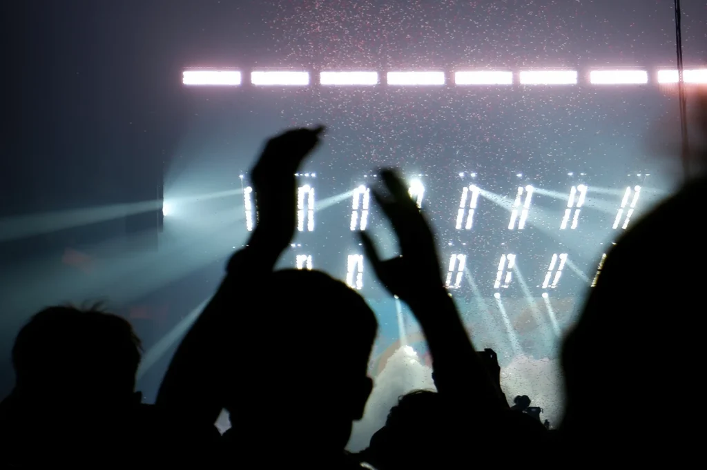 Silhouetted concert crowd cheering with raised hands under bright stage lights and atmospheric smoke during a live music performance in a dark indoor arena setting