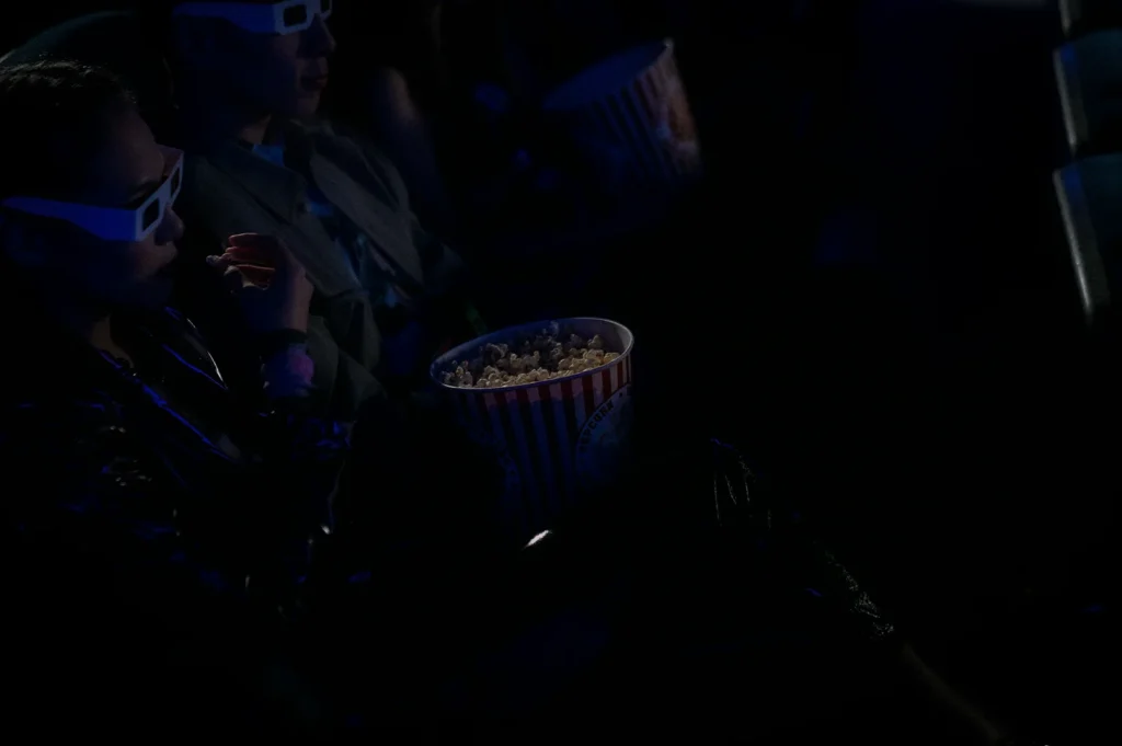 Audience members wearing 3D glasses while watching a movie in a dark cinema theater with popcorn buckets and dramatic blue lighting from the screen