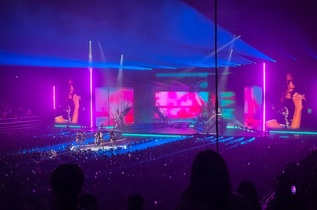Wide view of a Dua Lipa concert during the Radical Optimism Tour at Singapore Indoor Stadium, featuring a large stage with colorful LED screens, dramatic lighting, and a packed audience.