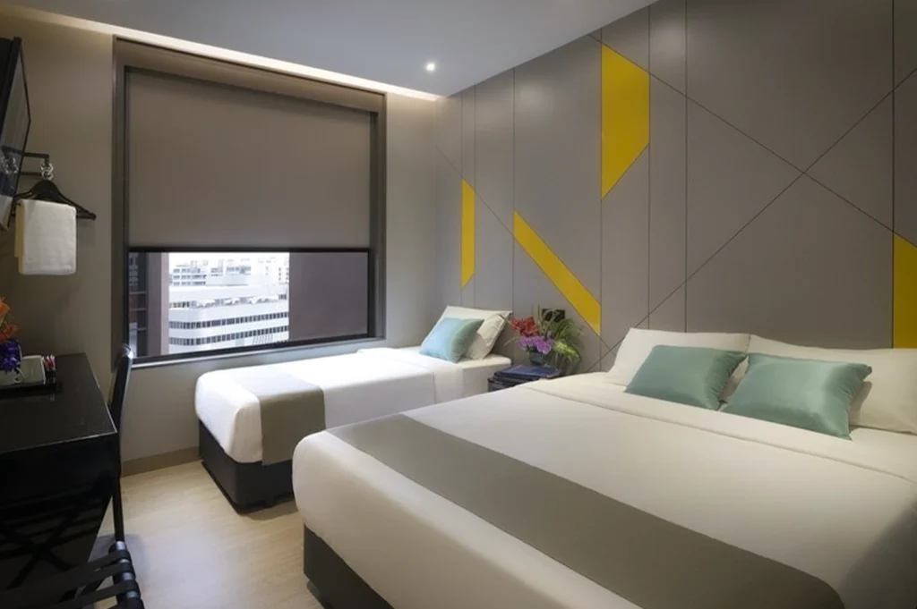 Modern twin room at Hotel Mi Bencoolen Singapore featuring two comfortable beds, minimalist grey and yellow wall design, large window with city view, and clean, compact contemporary interior.