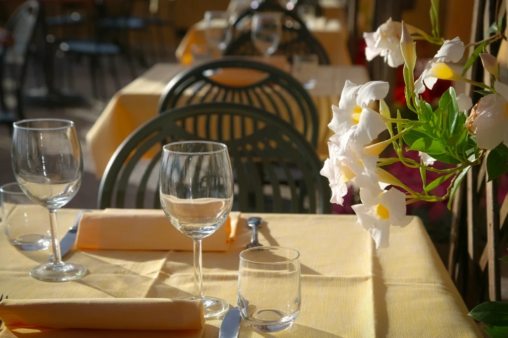 Outdoor restaurant table setting with wine glasses, napkins, and white flowers in sunlight, featuring elegant dining ambiance and cozy patio seating.