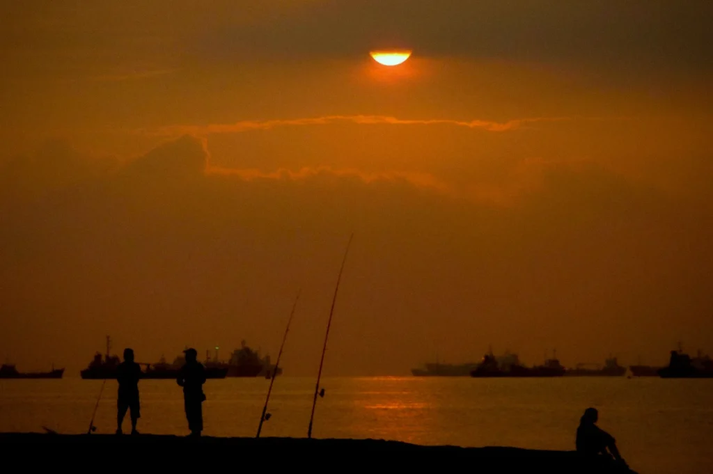 Golden sunrise over East Coast Park in Singapore with silhouettes of people fishing by the shoreline and ships resting on calm sea waters