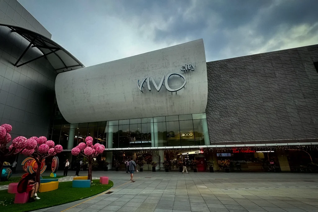 Exterior view of VivoCity shopping mall in Singapore with modern architecture, open plaza space, and decorative pink tree installations under a cloudy sky