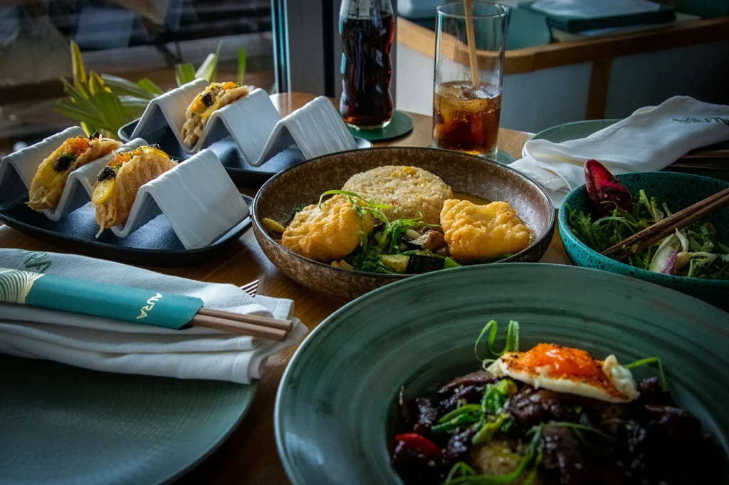 Asian fusion restaurant food spread in Singapore featuring gourmet tacos, fried rice, stir-fried beef bowl, fresh salad, and iced drinks served on a wooden dining table in a modern restaurant setting