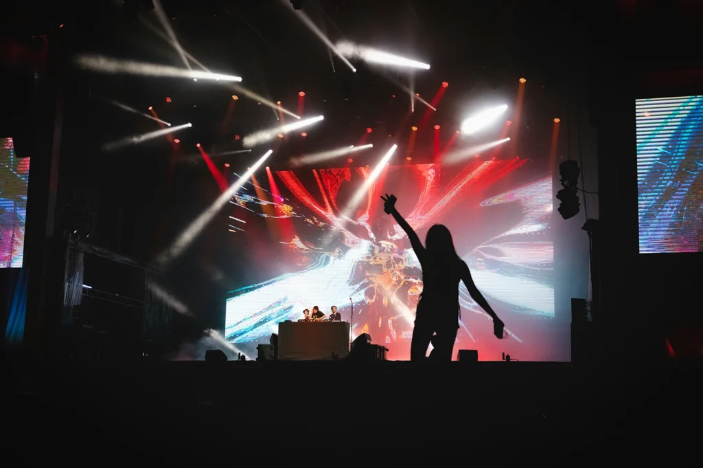 Silhouette of a person dancing in front of a DJ stage with bright concert lights and colorful LED visuals in a live music venue.