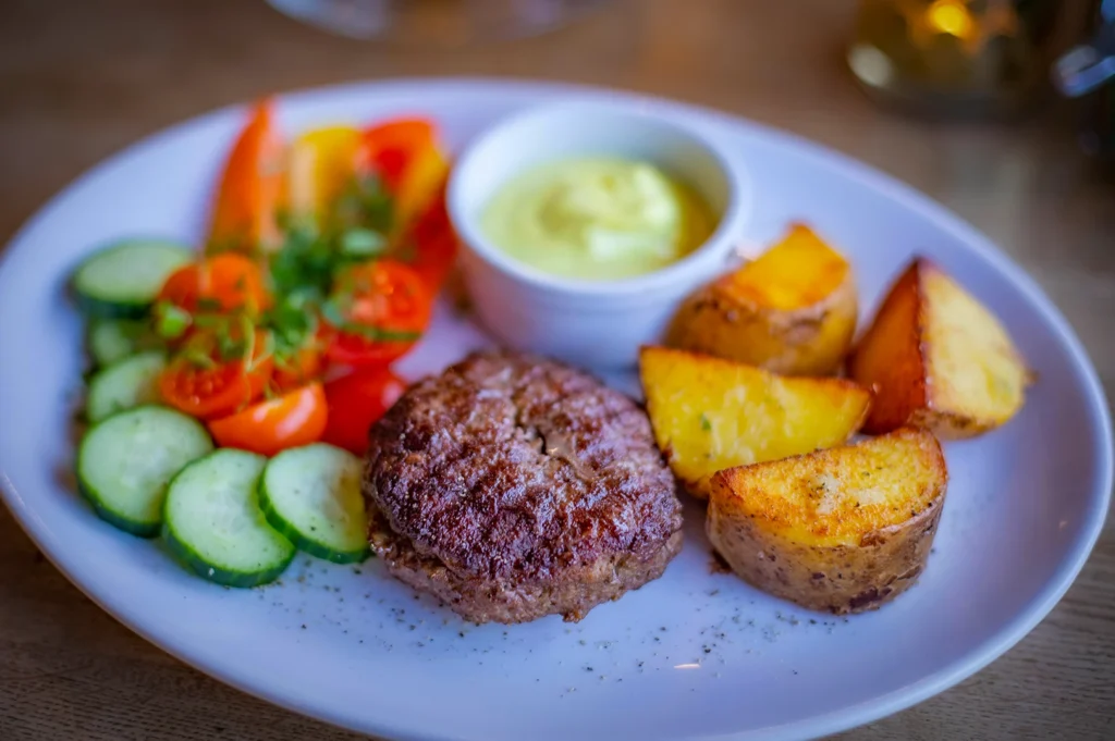 Grilled beef patty served with crispy roasted potato wedges fresh cucumber and tomato salad with creamy dipping sauce on a white plate at a casual restaurant dining table