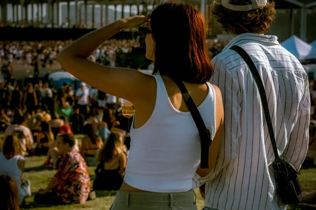 Two people standing and watching a large outdoor music festival crowd during a daytime concert event on a grassy field