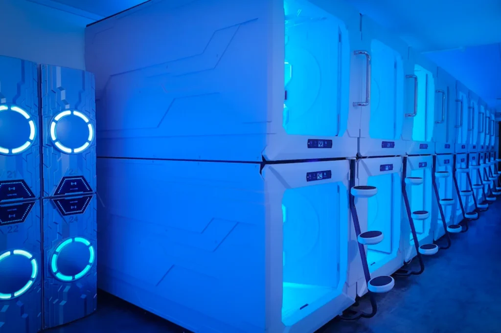 The photo shows the interior of a Capsule Pod Boutique Hostel at Boat Quay. It features a row of sleek, futuristic white sleeping capsules stacked two-high. Each pod has its own privacy screen or door, and the room is accented with blue, ambient LED lighting.