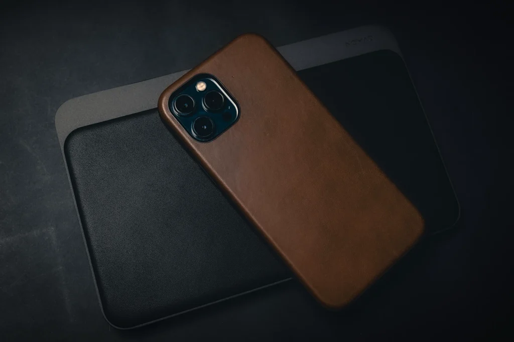 Apple iPhone with a brown leather case placed on top of a black leather desk mat in a dark minimalist workspace setup with moody lighting and premium accessories aesthetic