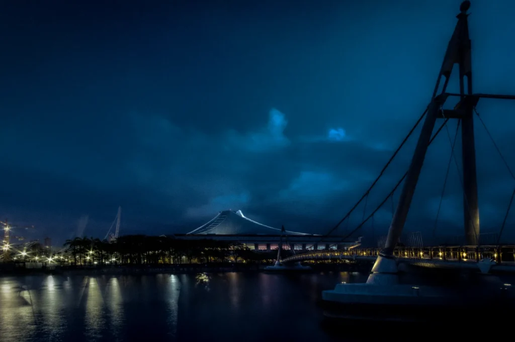 Night view of Kallang Indoor Stadium in Singapore with illuminated waterfront bridge, calm reflections on the water, and dramatic cloudy skies