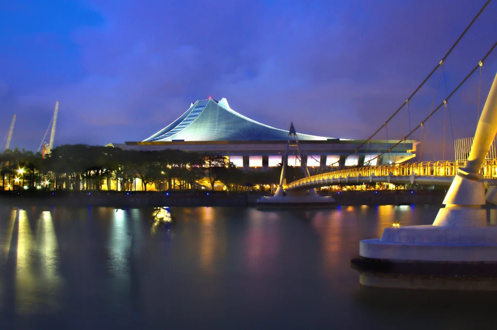 Night view of Singapore Indoor Stadium in Kallang with illuminated architecture, reflected lights on the water, and a pedestrian bridge in the foreground.