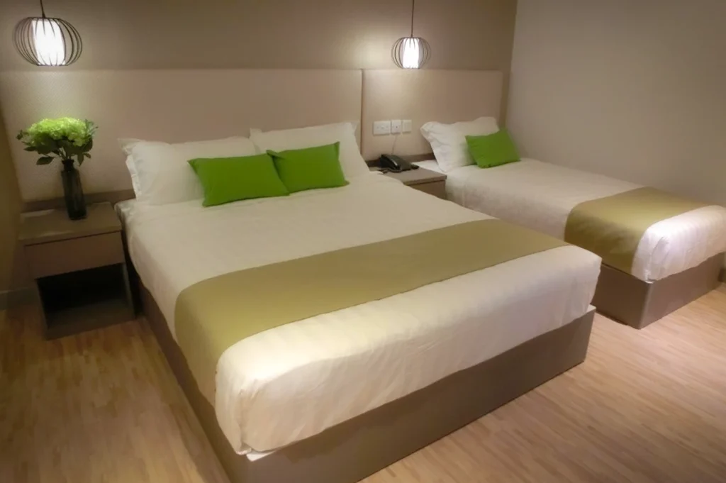 The guest room interior at Champion Hotel Joo Chiat in Singapore, featuring a clean and cozy setup with two beds. The larger bed is adorned with lime green accent pillows and a light olive bed runner, while a smaller single bed sits adjacent to it. Modern spherical pendant lights hang from the ceiling, casting a soft glow over the room's warm wood-textured flooring and minimalist decor.