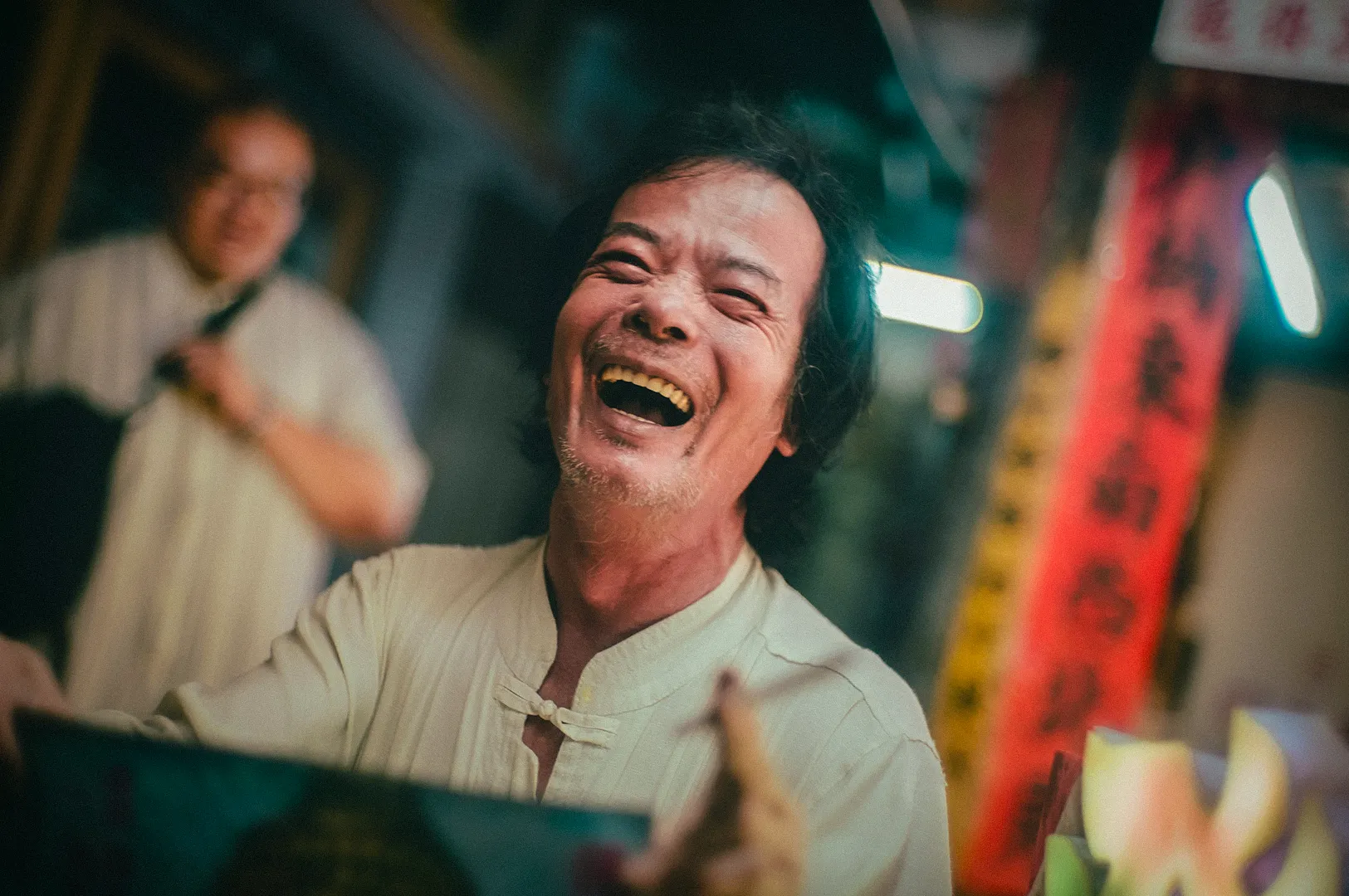 Laughing street vendor in a lively Asian market, captured in a candid moment with vibrant signage, warm lighting, and a bustling urban atmosphere in the background.