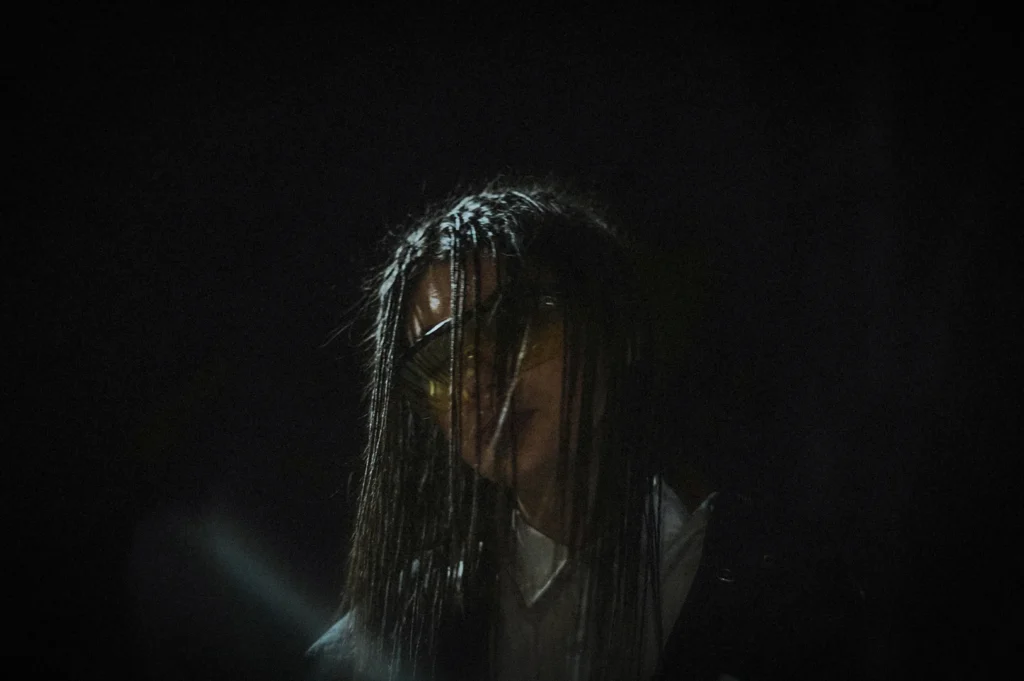 Creepy horror-themed figure with long dark hair partially covering the face, wearing reflective goggles in a dimly lit setting with dramatic shadows and an eerie atmosphere.