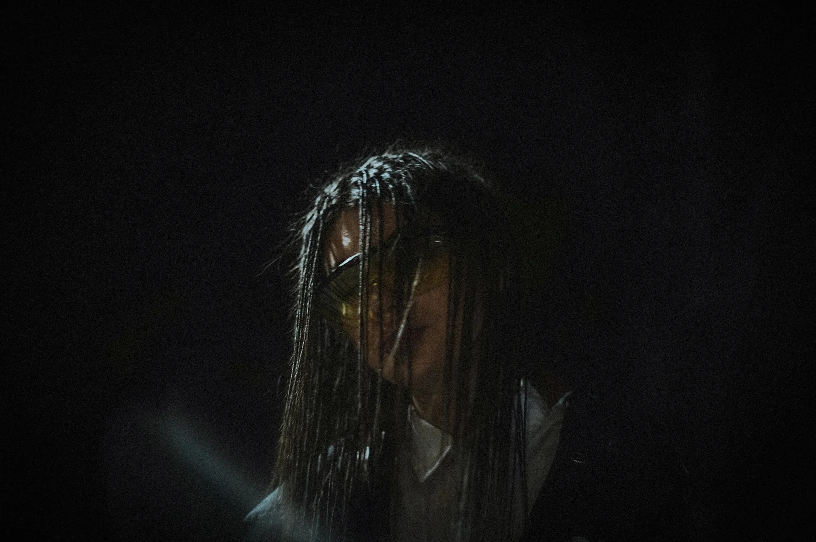 Creepy horror-themed figure with long dark hair partially covering the face, wearing reflective goggles in a dimly lit setting with dramatic shadows and an eerie atmosphere.