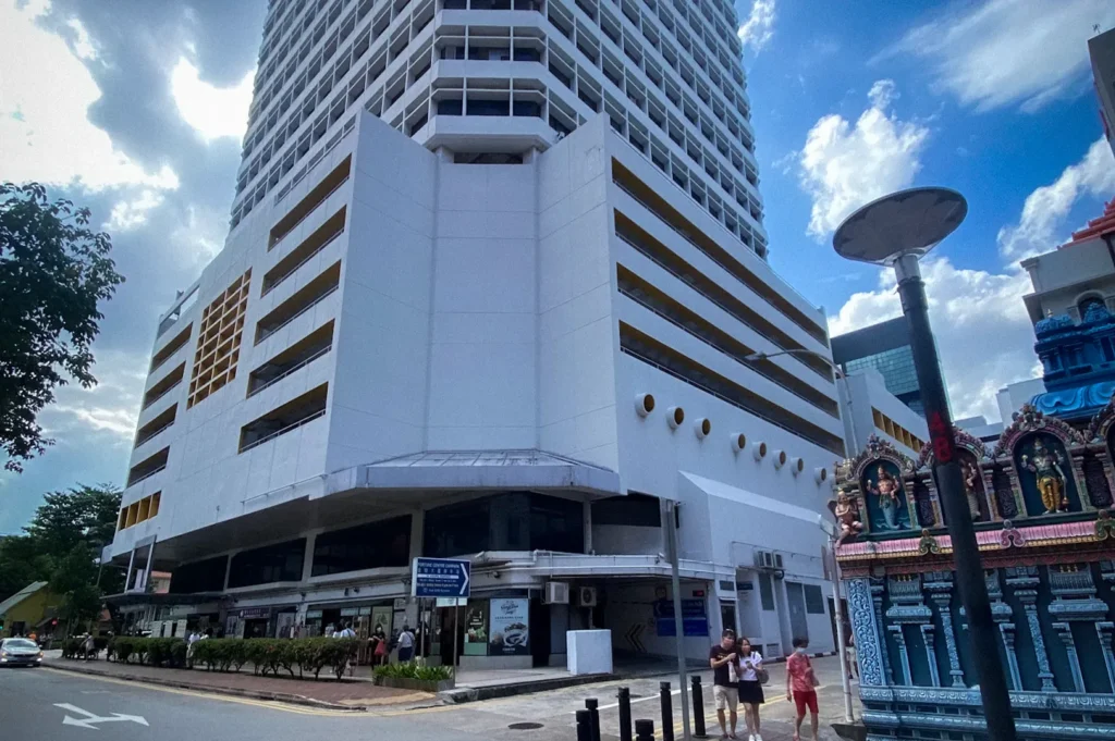 Exterior view of Fortune Centre Singapore, a prominent mixed-use commercial building with a white facade and yellow rooftop trim. The image shows the building towering above a street corner in the Bras Basah district, with a colorful Hindu temple in the foreground and pedestrians walking along the sidewalk under a partly cloudy sky.