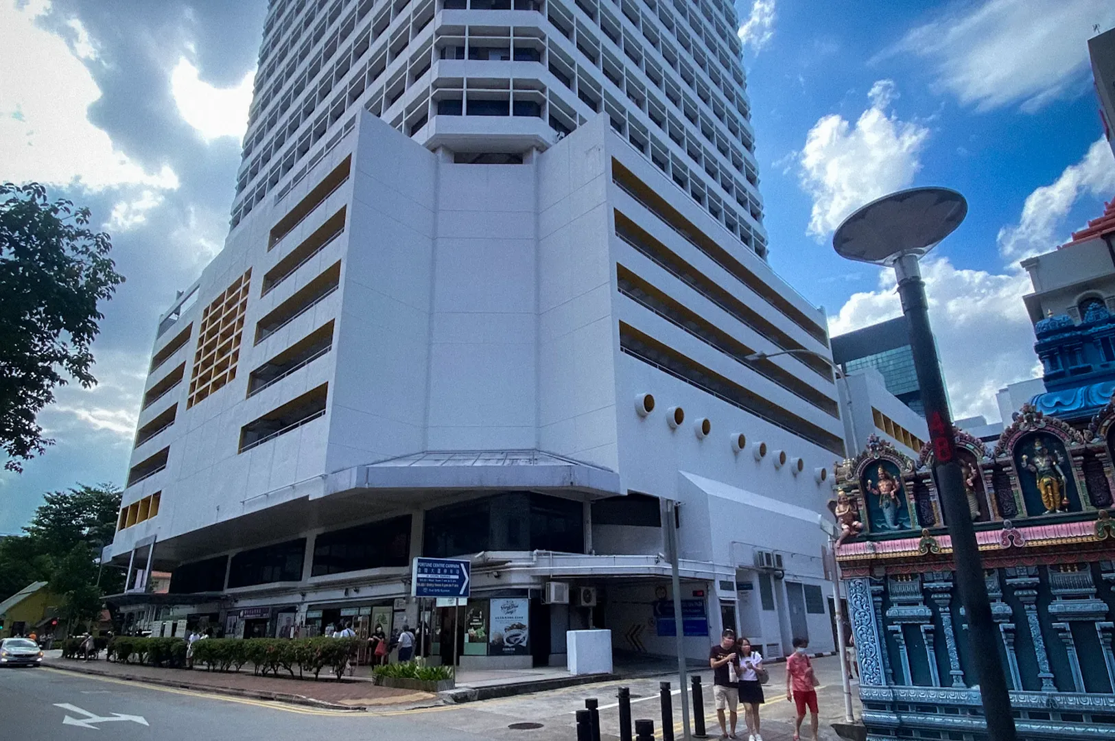 Exterior view of Fortune Centre Singapore, a prominent mixed-use commercial building with a white facade and yellow rooftop trim. The image shows the building towering above a street corner in the Bras Basah district, with a colorful Hindu temple in the foreground and pedestrians walking along the sidewalk under a partly cloudy sky.