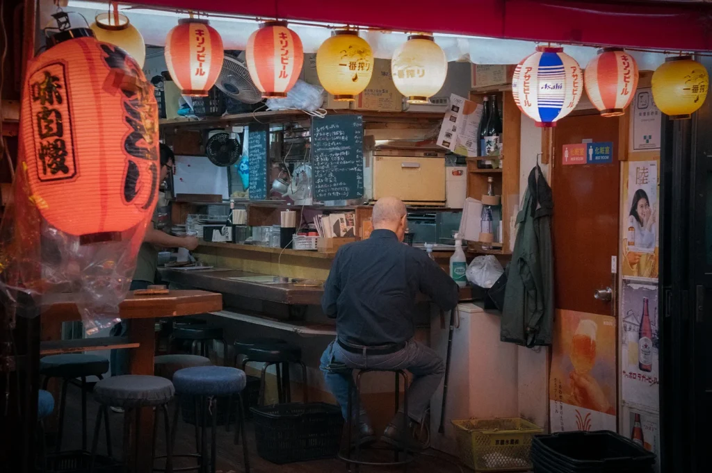 Traditional Japanese izakaya interior featuring illuminated paper lanterns, counter seating, wooden furnishings, handwritten menu boards, and a diner enjoying food and drinks in a cozy bar setting.