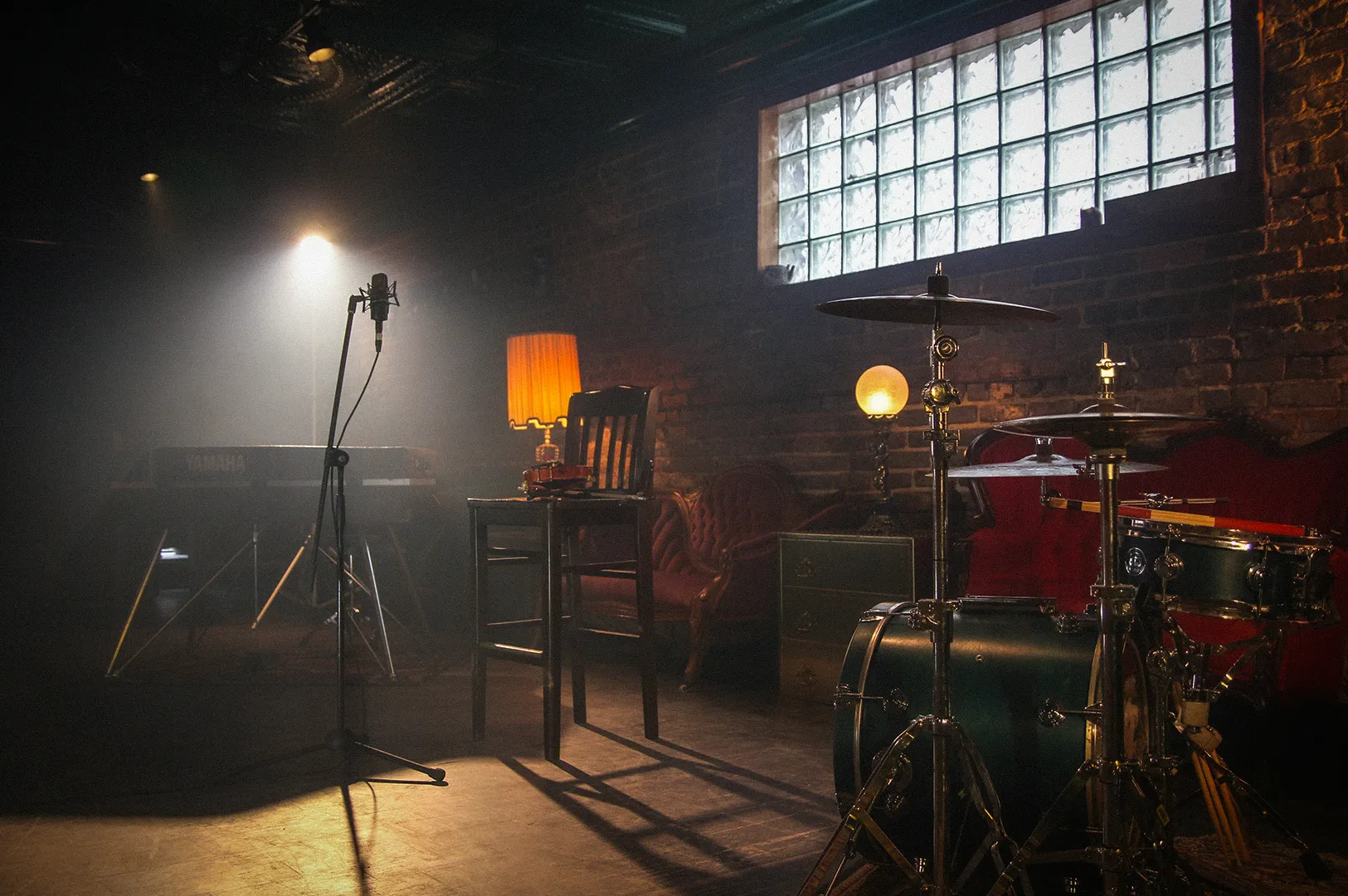 Cozy live music performance space featuring a drum set, microphone stand, keyboard, and vintage-style seating in a dimly lit brick-walled interior. Warm ambient lighting and stage equipment create an intimate atmosphere for acoustic sessions, jazz performances, and small live entertainment events.
